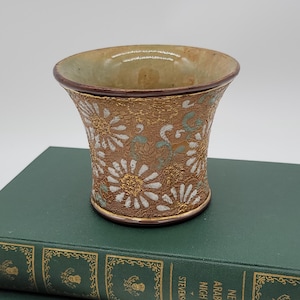 May include: A small, brown ceramic cup with a flared rim and a white and gold floral design. The cup is sitting on top of a stack of green books.