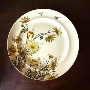 May include: A round, off-white ceramic plate featuring a floral design of yellow daisies and green stems. Two small blue bees are painted near the rim. The plate has a slightly aged appearance.