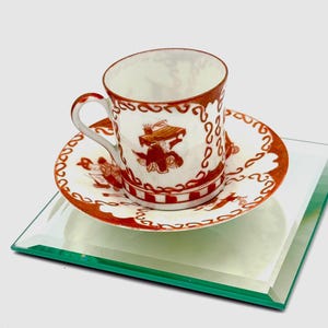 May include: A vintage teacup and saucer set. The cup and saucer are white with a red and white design. The cup has a handle and is sitting on the saucer. The set is on a mirrored surface.