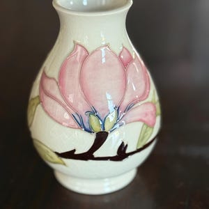 May include: Cream-colored ceramic vase with a pink and green floral design. The vase features a large pink flower with green leaves and a brown stem. The vase is approximately 6 inches tall.