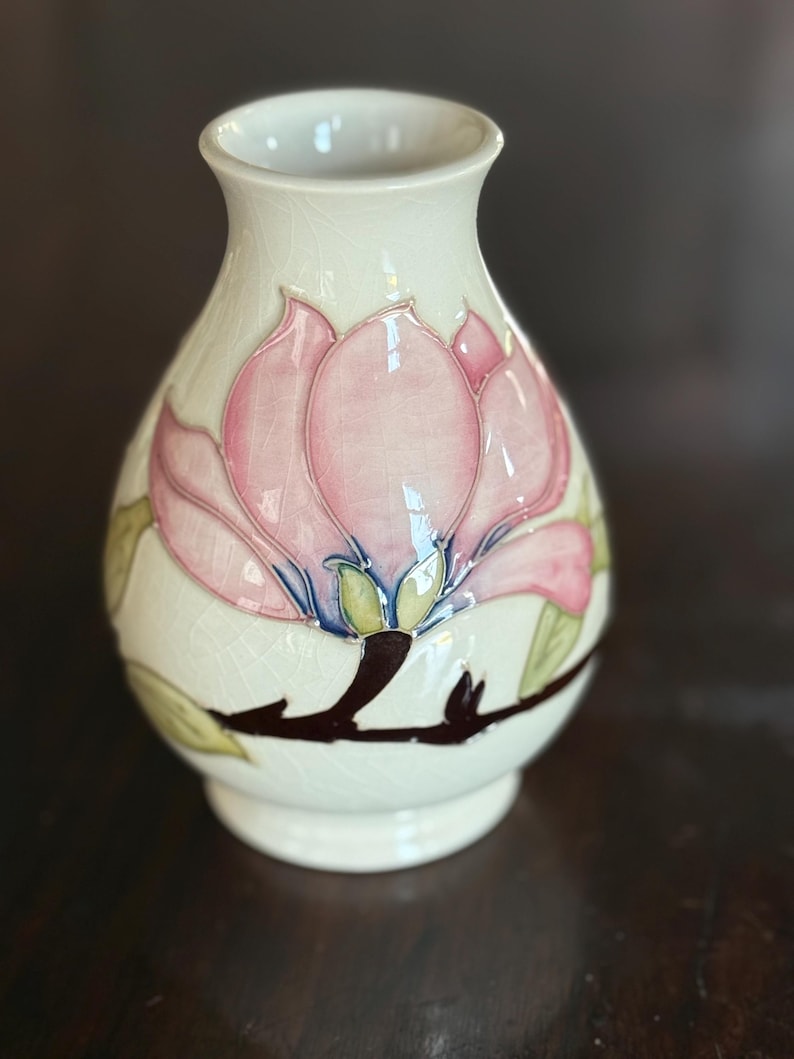 May include: Cream-colored ceramic vase with a pink and green floral design. The vase features a large pink flower with green leaves and a brown stem. The vase is approximately 6 inches tall.