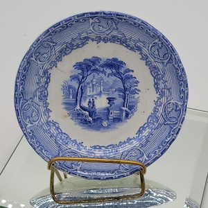 May include: A blue and white porcelain plate with a decorative border and a central image of a landscape with trees and a building. The plate is on a gold metal stand.