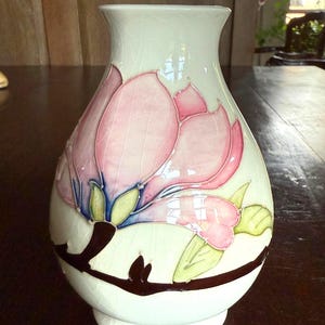 May include: A white ceramic vase with a pink floral design. The vase features a large pink flower with green and blue accents, and a brown branch. The vase has a wide base and a slightly flared top.