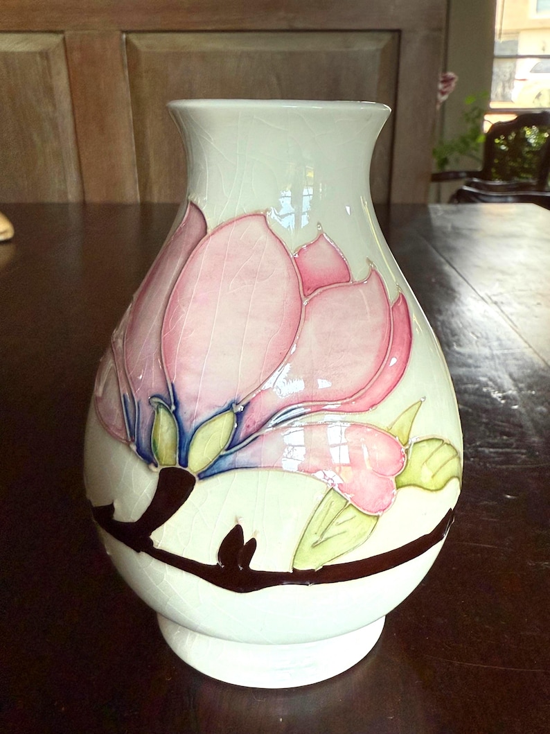 May include: A white ceramic vase with a pink floral design. The vase features a large pink flower with green and blue accents, and a brown branch. The vase has a wide base and a slightly flared top.