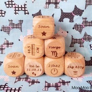 Peut inclure: Six cubes en bois avec des détails gravés, dont un nom, une date de naissance, une heure, un poids, une taille et un signe du zodiaque. Les cubes sont empilés en forme de pyramide sur un tissu à motifs bleu et blanc.