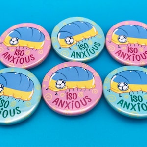 Puede incluir: Seis botones redondos y coloridos con un bicho bola de dibujos animados en cada uno. Los bichos bola son amarillos y azules con un ojo negro. Los botones tienen un fondo blanco con un patrón de remolino amarillo y el texto "iso anxious" en negro.