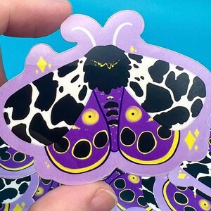 Peut inclure: Un autocollant représentant un papillon de nuit violet et noir avec des taches blanches. Le papillon a des yeux jaunes et une tête noire. L'autocollant est sur un fond violet avec une bordure blanche.