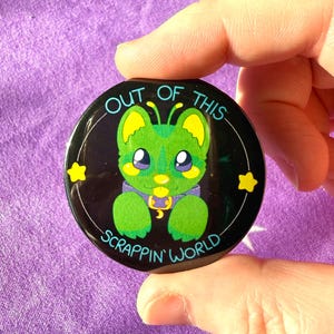 May include: A circular badge featuring a green alien character with large eyes and a purple collar. The text "OUT OF THIS SCRAPPIN' WORLD" is displayed around the alien on a black background.