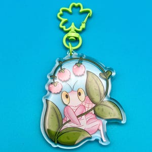 May include: A pink praying mantis with large, yellow eyes is surrounded by green leaves and pink flowers. The mantis is on a clear acrylic keychain with a green clover-shaped clip.