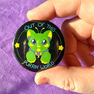 May include: A circular black badge featuring a green cartoon cat with yellow details and blue eyes, wearing a purple collar. The text "OUT OF THIS PURRIN' WORLD" surrounds the cat, with two yellow stars. The badge is held in a hand.