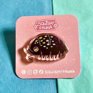 Può includere: Uno spillo a forma di porcellino di terra nero e bianco con accenti viola. Lo spillo è realizzato in acrilico trasparente e ha una finitura lucida. Lo spillo è su uno sfondo rosa con stelle bianche e il testo "SQUISHY FAUNA".