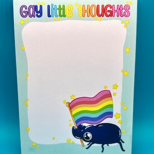 Op de afbeelding: Een lichtblauw notitieboekje met de tekst "Gay Little Thoughts" in regenboogkleuren. Het notitieboekje heeft een witte achtergrond met gele sterren en een cartoonkever die een regenboogvlag vasthoudt.