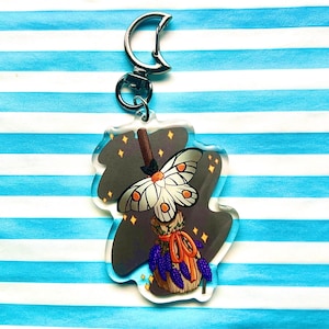 Peut inclure: Un porte-clés en acrylique transparent avec une illustration fantaisiste d'un balai sur lequel est perché un papillon de nuit. Le balai est décoré de fleurs violettes et d'un ruban rouge. L'arrière-plan est gris foncé avec des étoiles dorées.