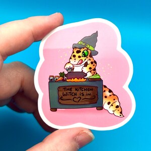 Può includere: Un adesivo rosa con un geco dei cartoni animati che indossa un cappello da strega e cucina in una pentola. Il testo sull'adesivo dice "The kitchen witch is in".