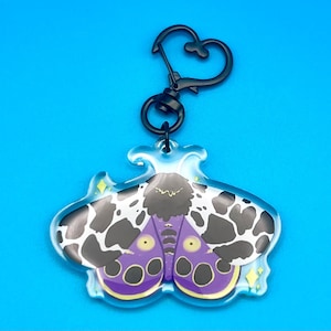 May include: A clear acrylic keychain featuring a moth design with black and white spots, purple accents, and yellow details. The keychain has a black heart-shaped clasp and a black ring. The background is a solid blue.