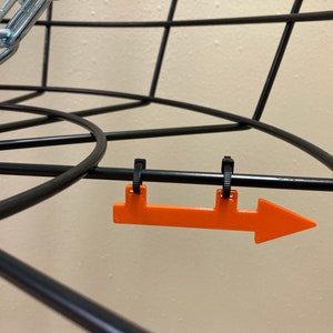 May include: Close-up of a black disc golf basket with an orange arrow attached to the top bar. The arrow is pointing to the right.