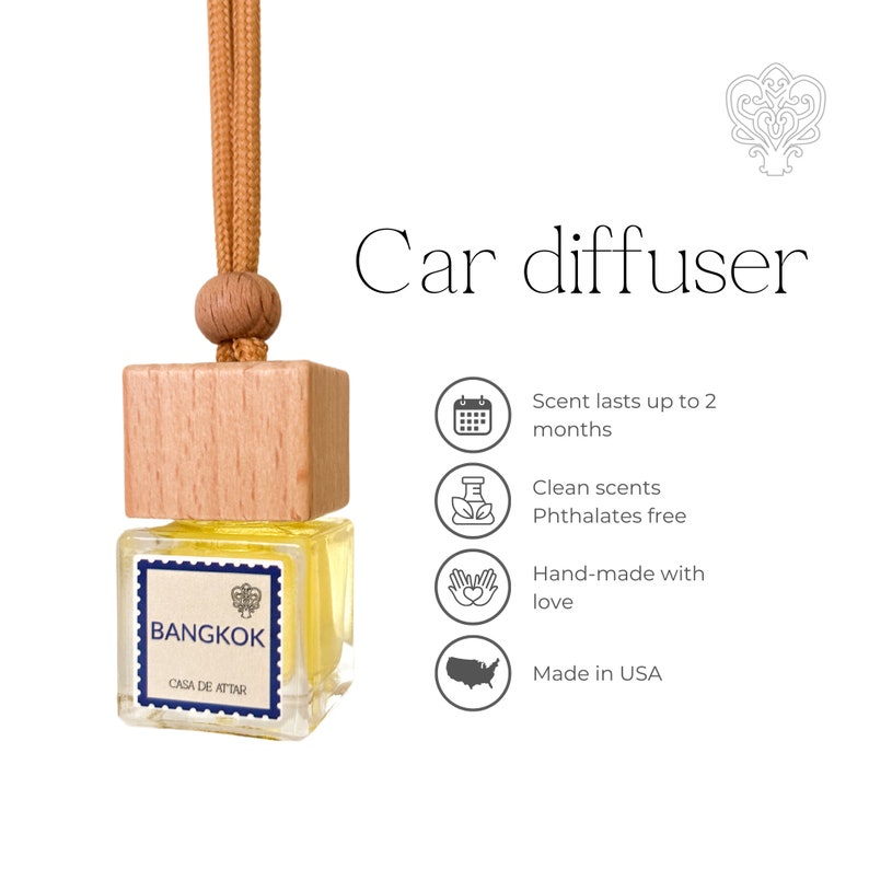 Op de afbeelding: Een houten autodiffuser met een gele vloeistof erin. De diffuser heeft een houten dop en een touwtje om op te hangen. Het label op de diffuser zegt "BANGKOK CASA DE ATTAR". De afbeelding bevat ook pictogrammen voor de geurduur, schone geuren, handgemaakt met liefde en gemaakt in de VS.