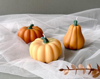 May include: Three small pumpkin-shaped candles on a white tulle fabric. The candles are in shades of orange and yellow.