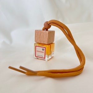 Op de afbeelding: Een klein, doorzichtig glazen flesje met een houten dop en een geel touwtje. Het flesje is gevuld met een gele vloeistof en heeft een etiket met de tekst "Positano Casa de Attar".