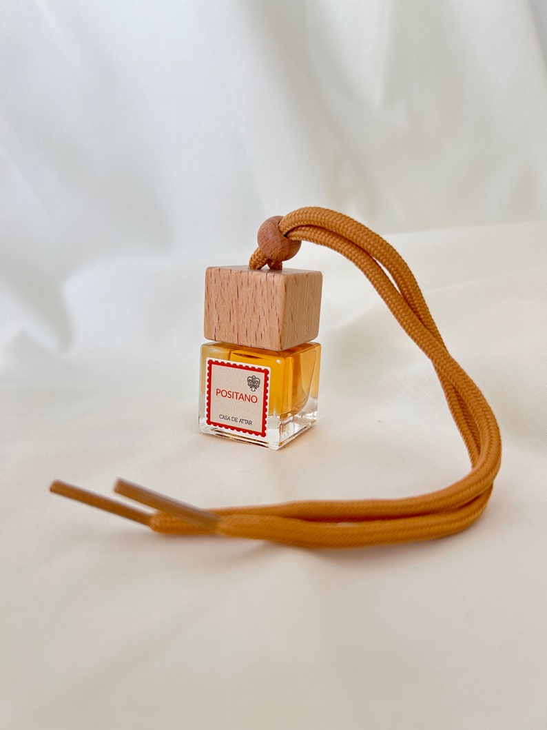 Op de afbeelding: Een klein, doorzichtig glazen flesje met een houten dop en een geel touwtje. Het flesje is gevuld met een gele vloeistof en heeft een etiket met de tekst "Positano Casa de Attar".