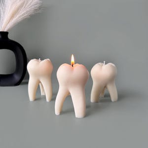 May include: Three white tooth-shaped candles, one with a lit wick, on a gray surface.