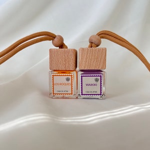 Op de afbeelding: Twee kleine glazen flesjes met houten dopjes en bruine koordhangers. De flesjes bevatten oranje en paarse vloeistoffen. De etiketten lezen "Los Roques" en "Waikiki" met de tekst "Casa de Attar" eronder.
