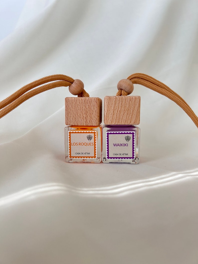 Op de afbeelding: Twee kleine glazen flesjes met houten dopjes en bruine koordhangers. De flesjes bevatten oranje en paarse vloeistoffen. De etiketten lezen "Los Roques" en "Waikiki" met de tekst "Casa de Attar" eronder.