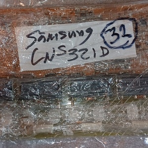 May include: A close-up of a Samsung TV circuit board wrapped in plastic. A white label with black writing reads "Samsung 32" and "CN-321D".