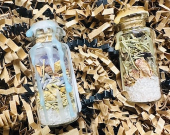 May include: Two small glass bottles filled with dried herbs and crystals. The bottles are sealed with cork stoppers and have a wax seal on top. The bottles are sitting on a bed of brown packing peanuts.
