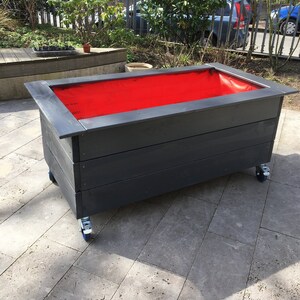 Könnte beinhalten: Ein schwarzer Holz-Pflanzkasten mit roter Auskleidung und Rädern. Der Kasten steht auf einer grauen Terrasse.