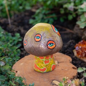 May include: A whimsical, hand-painted mushroom sculpture with a brown cap adorned with multiple painted eyes. The stem features a yellow and orange pattern. The sculpture is displayed on a wooden slice, surrounded by greenery.