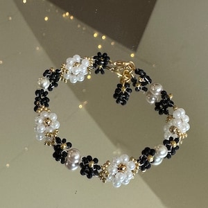 May include: A beaded bracelet featuring a floral design. The bracelet is composed of white and black beads, with gold accents. The clasp is gold-toned. The bracelet is designed with a flower pattern.