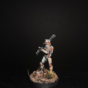 Puede incluir: Una miniatura pintada de un soldado clon de Star Wars, con armadura blanca y naranja, sosteniendo un rifle bláster. La figura está de pie sobre una pequeña base marrón con hierba verde.