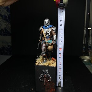 May include: A painted miniature figure of a Mandalorian warrior from the Star Wars universe. The figure is standing on a base with a Mandalorian symbol. The figure is approximately 10 inches tall.