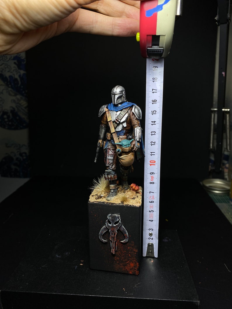 May include: A painted miniature figure of a Mandalorian warrior from the Star Wars universe. The figure is standing on a base with a Mandalorian symbol. The figure is approximately 10 inches tall.