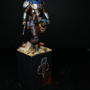 May include: A painted miniature figure of a Mandalorian warrior from the Star Wars universe, holding a small green alien figure. The figure is standing on a base with a Mandalorian symbol painted on it.