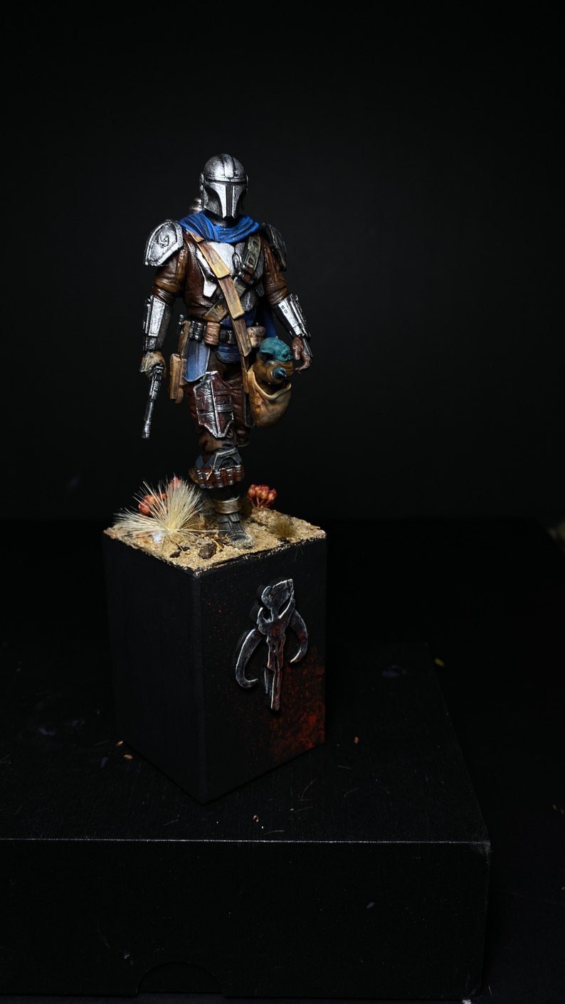 May include: A painted miniature figure of a Mandalorian warrior from the Star Wars universe, holding a small green alien figure. The figure is standing on a base with a Mandalorian symbol painted on it.