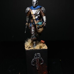 May include: A painted action figure of a Mandalorian warrior, wearing a silver and blue helmet and armor, holding a small green creature in a bag. The figure is standing on a black base with a silver Mandalorian symbol.