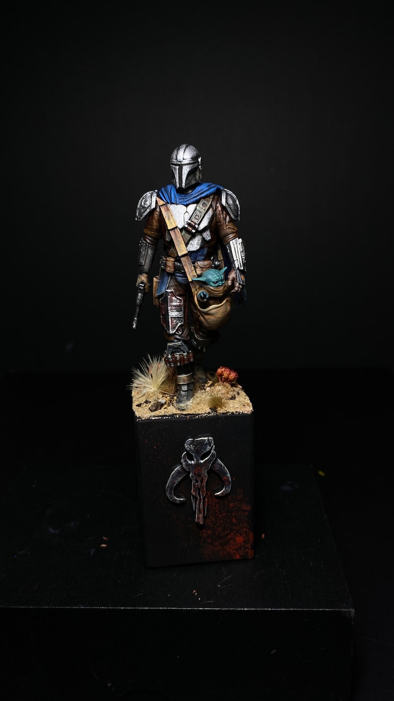 May include: A painted action figure of a Mandalorian warrior, wearing a silver and blue helmet and armor, holding a small green creature in a bag. The figure is standing on a black base with a silver Mandalorian symbol.