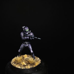 May include: A black and purple painted action figure of a Star Wars clone trooper holding a blaster rifle. The figure is standing on a small, sandy base.