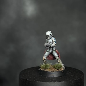 Op de afbeelding: Miniatuurfiguur van een soldaat in witte en zilveren bepantsering, poserend met een wapen. De figuur heeft een rode cape en staat op een kleine, ronde basis met gras. De achtergrond is een donkere, vage gradiënt.