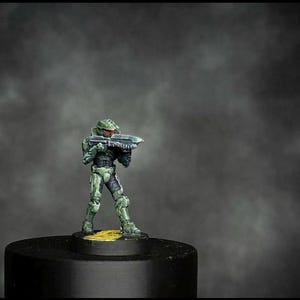 May include: A small figurine of a soldier in green and black armour, holding a futuristic rifle. The figure stands on a black base against a dark, smoky background. The soldier's helmet and armour have detailed features.