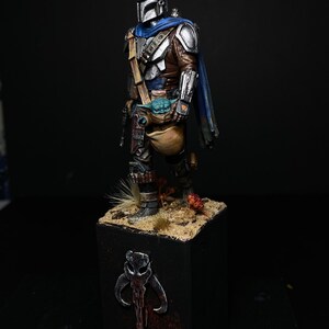 May include: A painted miniature figurine of a Mandalorian warrior from the Star Wars universe. The figure is wearing a silver and blue armor with a brown leather belt and a blue cape. The Mandalorian is holding a small green alien creature in his right hand. The figure is standing on a black base with a Mandalorian symbol.