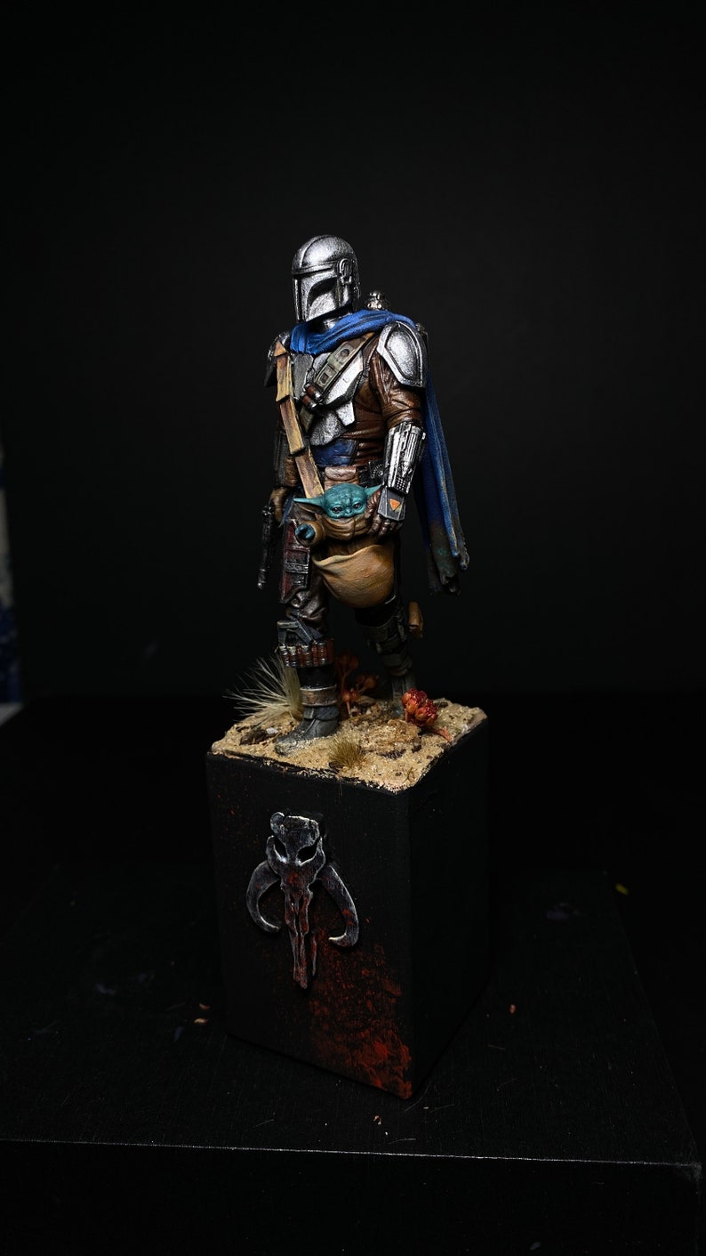 May include: A painted miniature figurine of a Mandalorian warrior from the Star Wars universe. The figure is wearing a silver and blue armor with a brown leather belt and a blue cape. The Mandalorian is holding a small green alien creature in his right hand. The figure is standing on a black base with a Mandalorian symbol.