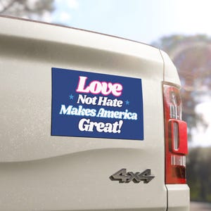 May include: A rectangular blue bumper sticker with the words "Love Not Hate Makes America Great!" in white and pink lettering is affixed to the back of a white vehicle. The sticker has a star on each side of the words "Not Hate".