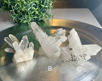 May include: Three clear quartz crystal clusters with small, dark inclusions are displayed on a silver tray. The crystals vary in size and shape, with some having multiple points. The crystals are labeled A, B, and C.