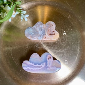 May include: Two cloud-shaped polished stones with unique patterns and colours. Stone A is a mix of beige, white, and pink hues, while Stone B features shades of purple, blue, and white. Both are displayed on a gold-coloured surface.