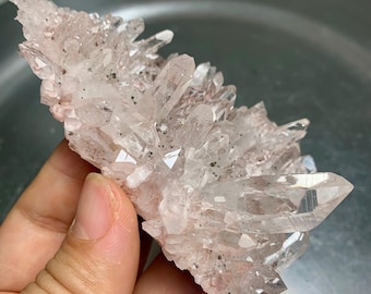 May include: A cluster of clear quartz crystals with a pale pink hue and small black inclusions. The crystals are pointed and clustered together, creating a textured appearance. The quartz is held in a hand.