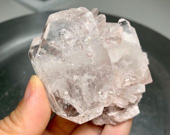 May include: A cluster of clear, translucent quartz crystals with a pale pink hue. The crystals have sharp, geometric shapes and are held in a hand. The overall appearance is natural and mineral-like.