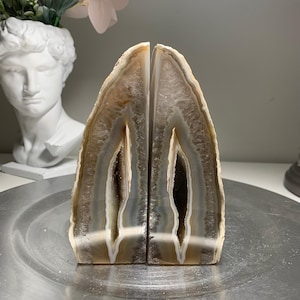 May include: Two agate bookends with a natural, layered appearance. The bookends feature a mix of beige, grey, and white tones, with a polished surface and a hollowed-out centre. These decorative bookends add a touch of natural elegance to any bookshelf.